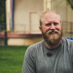 A cheerful bearded man smiling outdoors in a casual setting with greenery.